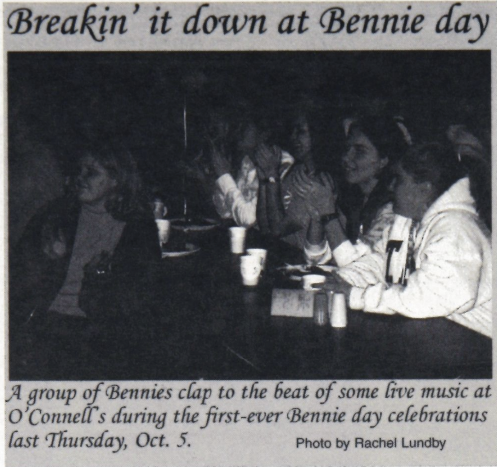 A group of people sit at a table clapping to the beat of live music at O’Connell’s during the first-ever Bennie Day celebrations. The scene features dim lighting, and the photo is credited to Rachel Lundby.